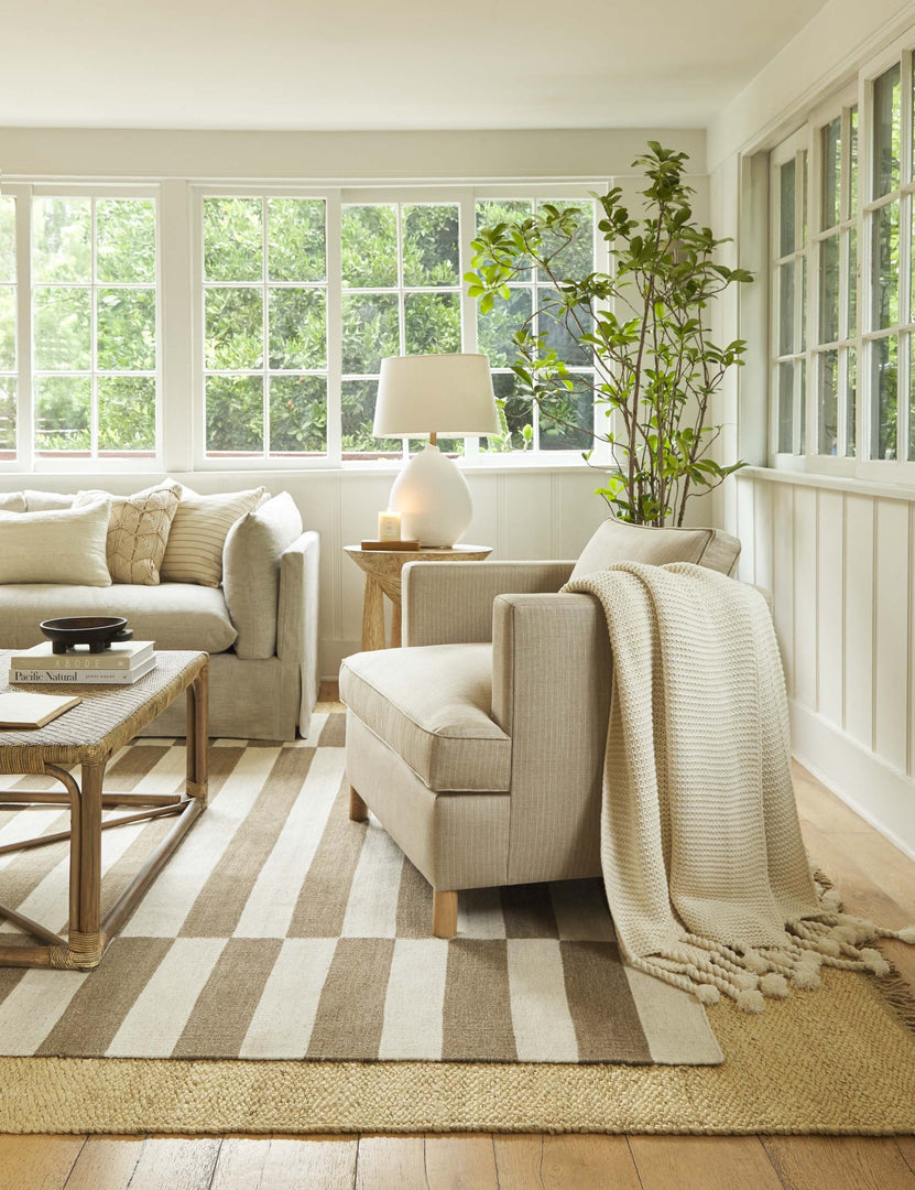 #color::stripe | The Belmont stripe linen accent chair sits atop two layered rugs in a room with wrap-around windows and a woven coffee table