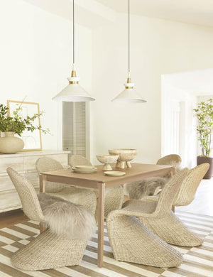 The Kloe conical Pendant Light with a white finish and aged brass hardware is mounted in a dining room with neutral woven dining chairs, a light wooden dining table, and a white and brown striped rug
