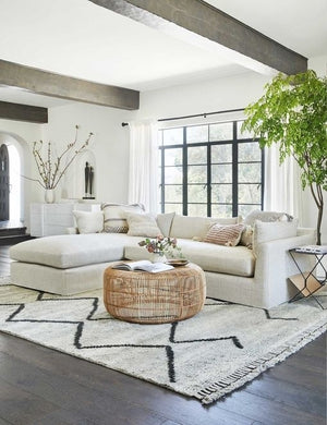 The Amina round coffee table sits atop a gray patterned rug in front of a natural linen sectional sofa