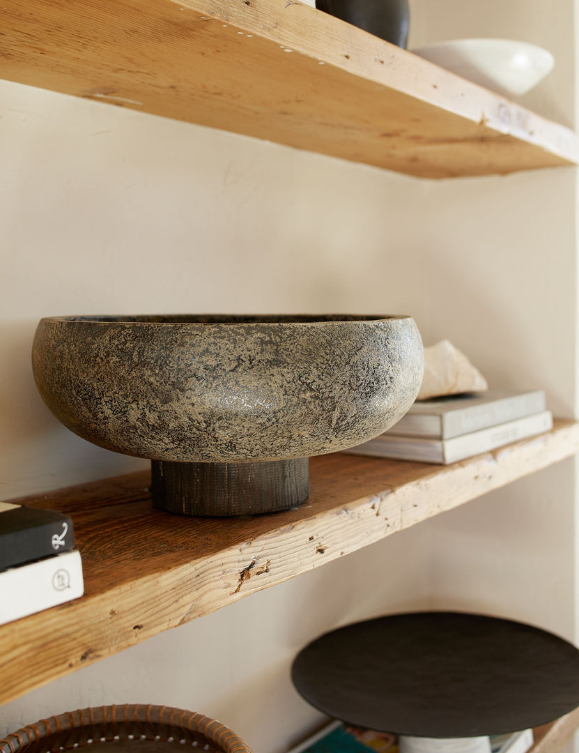 | Close-up of the Lakshmi terracotta bowl with antiqued black finish and woven base sitting on a wooden shelf with other centerpiece decor and stacks of books