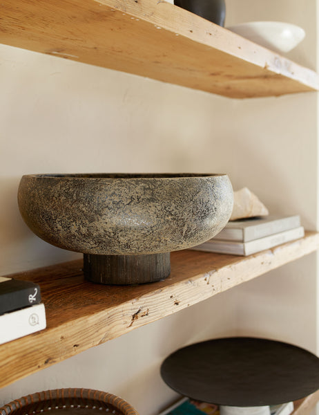 | Close-up of the Lakshmi terracotta bowl with antiqued black finish and woven base sitting on a wooden shelf with other centerpiece decor and stacks of books