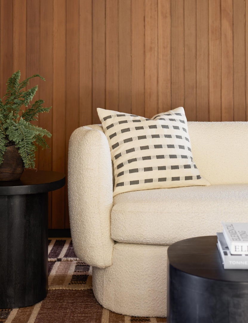 #color::pumice | Bertu pumice gray pillow with a woven dash pattern by Bolé Road Textiles sits on a cream boucle rounded sofa in a room with wooden paneled walls