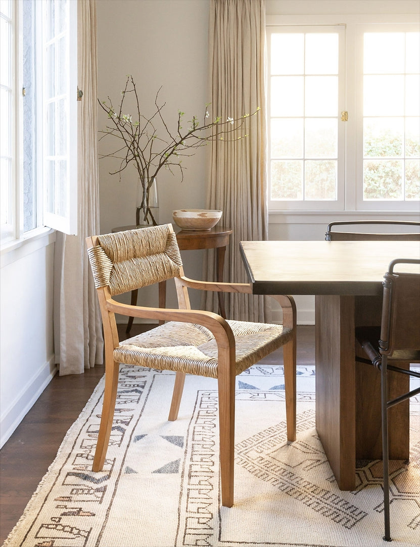 | The Nolani woven rattan arm chair is sitting in a dining room at the head of a wooden rectangular dining table atop a geometric patterned rug.