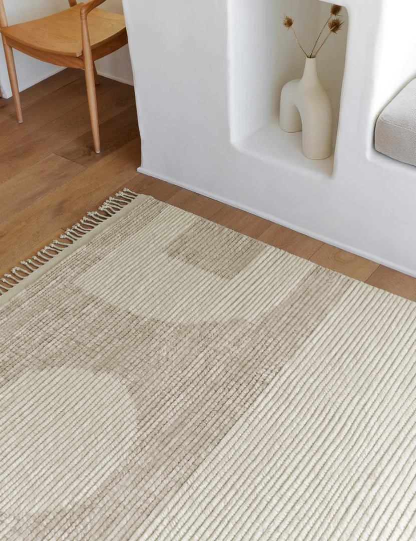 #size::2-6--x-8- #size::6--x-9- #size::8--x-10- #size::9--x-12- #size::10--x-14- #size::12--x-15- | Close-up of the Nomad neutral-toned geometric floor rug by Élan Byrd with subtle ribbed design in a room with a sculptural white vase and a neutral wooden dining table atop hardwood floors