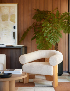 The Celeste honey wood accent chair with wishbone frame sits in a retro living room with wood paneled walls, a black planter, and a round wooden coffee table.