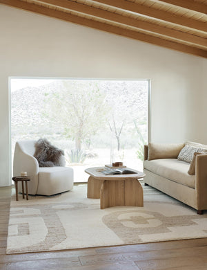The Oasis plush geometric neutral-toned rug by Élan Byrd lays in a living room with an oval wooden coffee table, a white linen accent chair, and a light brown velvet sofa