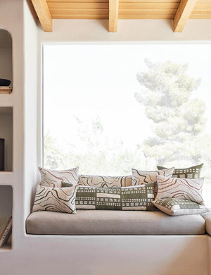 The Stonewalk green geometric throw pillow by Élan Byrd sits on a natural linen cushioned bench with other terracotta and green toned throw pillows in front of a large window