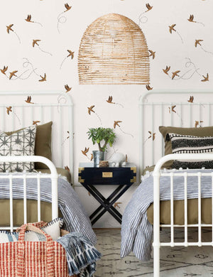 The Sparrow brown wallpaper by Rylee and Cru is in a bedroom with two white victorian style beds with a jute chandelier and black and gold bedside table in the center.