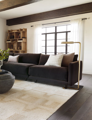 The Reid c-shape aged brass Floor Lamp sits in a living room with a brown velvet sofa, a sculptural wooden bookshelf, and a natural-toned plush rug
