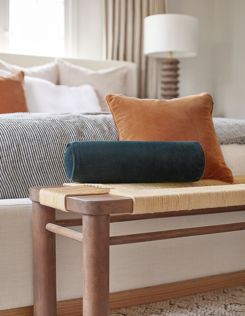 #color::navy | The Sabine navy velvet cylindrical bolster pillow sits atop a woven bench with a burnt orange velvet throw pillow in a bedroom at the end of a natural linen framed bed