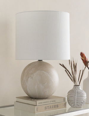 The Vivienne tan ceramic table lamp with spherical base sits atop a glass sideboard with a white vase and a stack of books